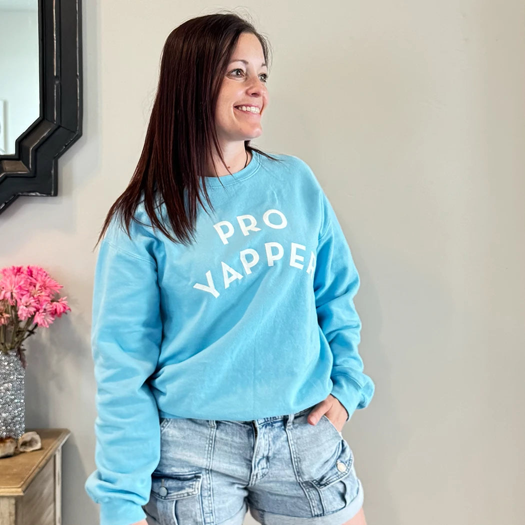 A fair-skinned person with shoulder-length dark hair smiles to their right, wearing a light blue crewneck sweatshirt with the words "PRO" stacked above "YAPPER" in a simple, rounded white font on the chest. graphic tee hoodies