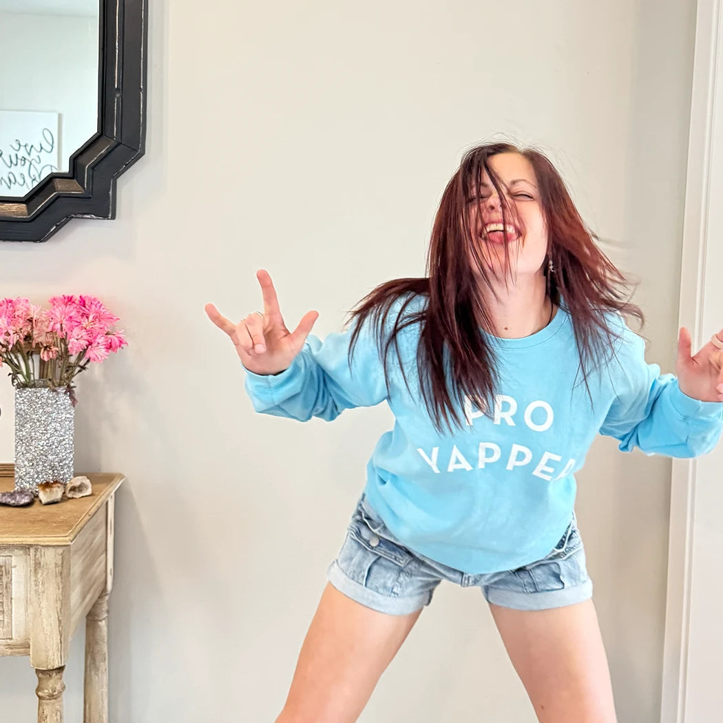 A fair-skinned person with shoulder-length dark hair smiles to their right, wearing a light blue crewneck sweatshirt with the words "PRO" stacked above "YAPPER" in a simple, rounded white font on the chest. graphic tee hoodies