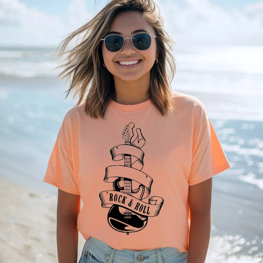 A smiling woman with shoulder-length blonde hair and sunglasses stands on a beach, wearing a peach-colored graphic apparel t-shirt with a black graphic of an electric guitar wrapped in a banner that reads "ROCK & ROLL." The ocean and a clear sky are visible in the background.