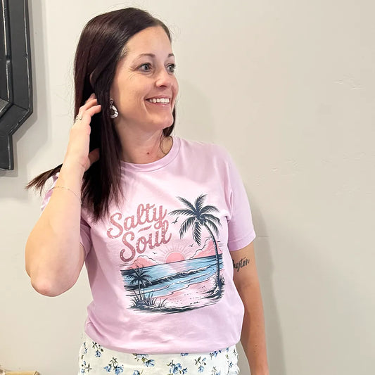 A fair-skinned person with shoulder-length dark hair smiles, wearing a light purple graphic apparel t-shirt with a beach-themed graphic. The graphic includes a sunset with pink and orange hues over a blue ocean, a silhouetted palm tree, and the cursive text "Salty Soul" arched above the scene.