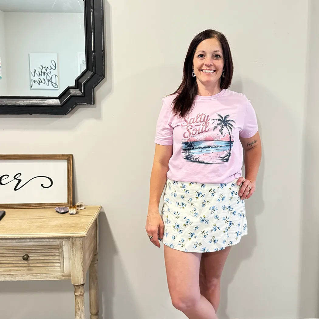 A fair-skinned person with shoulder-length dark hair smiles, wearing a light purple graphic apparel t-shirt with a beach-themed graphic. The graphic includes a sunset with pink and orange hues over a blue ocean, a silhouetted palm tree, and the cursive text "Salty Soul" arched above the scene.