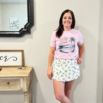A fair-skinned person with shoulder-length dark hair smiles, wearing a light purple graphic apparel t-shirt with a beach-themed graphic. The graphic includes a sunset with pink and orange hues over a blue ocean, a silhouetted palm tree, and the cursive text "Salty Soul" arched above the scene.
