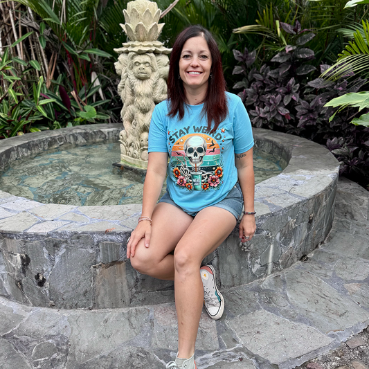 A smiling woman with dark hair stands on a stone path in a lush, green garden. She is wearing a light blue t-shirt with a "Stay Weird" graphic featuring a skeleton and flowers, and light wash denim shorts. She has her hands in her pockets and is looking directly at the camera. White sneakers complete her casual outfit.