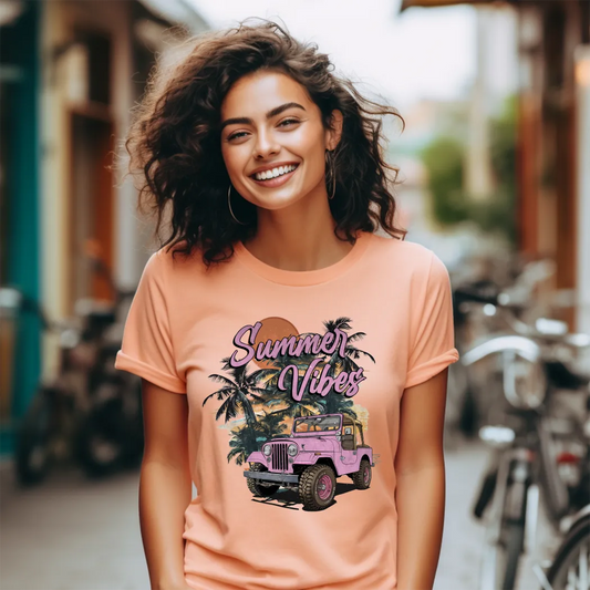 A smiling woman with curly brown hair is the focus of the image, wearing a light peach-colored graphic apparel t-shirt. The graphic apparel t-shirt features a vibrant graphic with the text "Summer Vibes" in a stylized pink script, layered over an illustration of palm trees, a setting sun in shades of orange and yellow, and a pink vintage jeep.