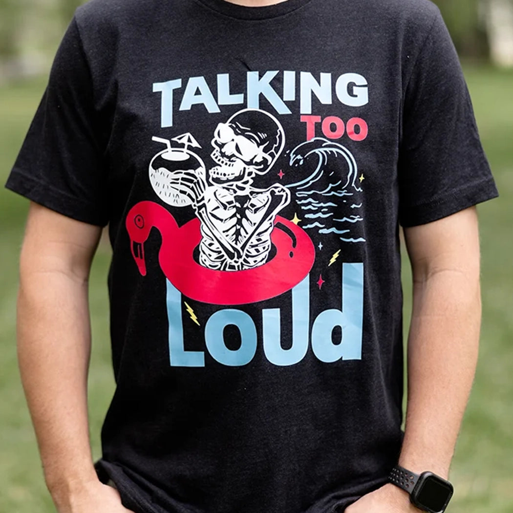 Black Talking Too Loud shirt with wave, skeleton in a pink flamingo floaty with a coconut drink. Design is blue, pink and white. 