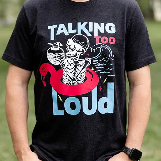 Black Talking Too Loud shirt with wave, skeleton in a pink flamingo floaty with a coconut drink. Design is blue, pink and white. 