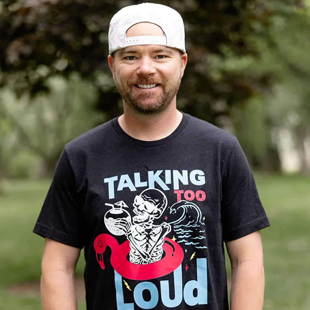 Black Talking Too Loud shirt with wave, skeleton in a pink flamingo floaty with a coconut drink. Design is blue, pink and white. 