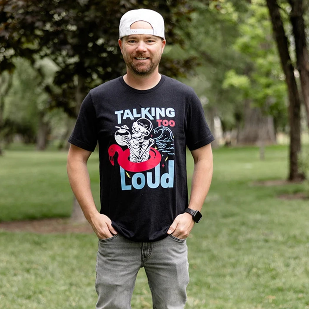 Black Talking Too Loud shirt with wave, skeleton in a pink flamingo floaty with a coconut drink. Design is blue, pink and white. 