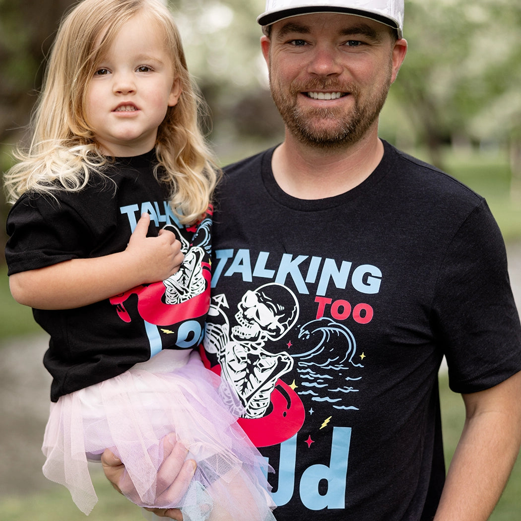 Black Talking Too Loud shirt with wave, skeleton in a pink flamingo floaty with a coconut drink. Design is blue, pink and white. 