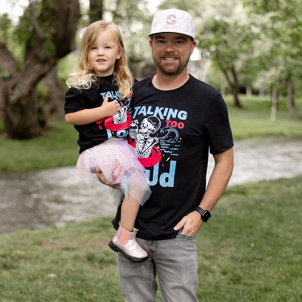 Black Talking Too Loud shirt with wave, skeleton in a pink flamingo floaty with a coconut drink. Design is blue, pink and white. 