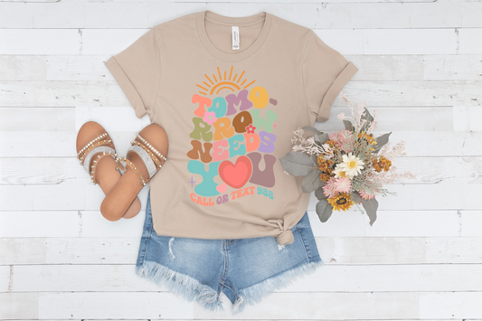 Tan shirt with shorts with sandals and flowers next to it. The shirt says Tomorrow Needs You Call or Text 988. The design has flowers a sunshine and is in bright colors. 