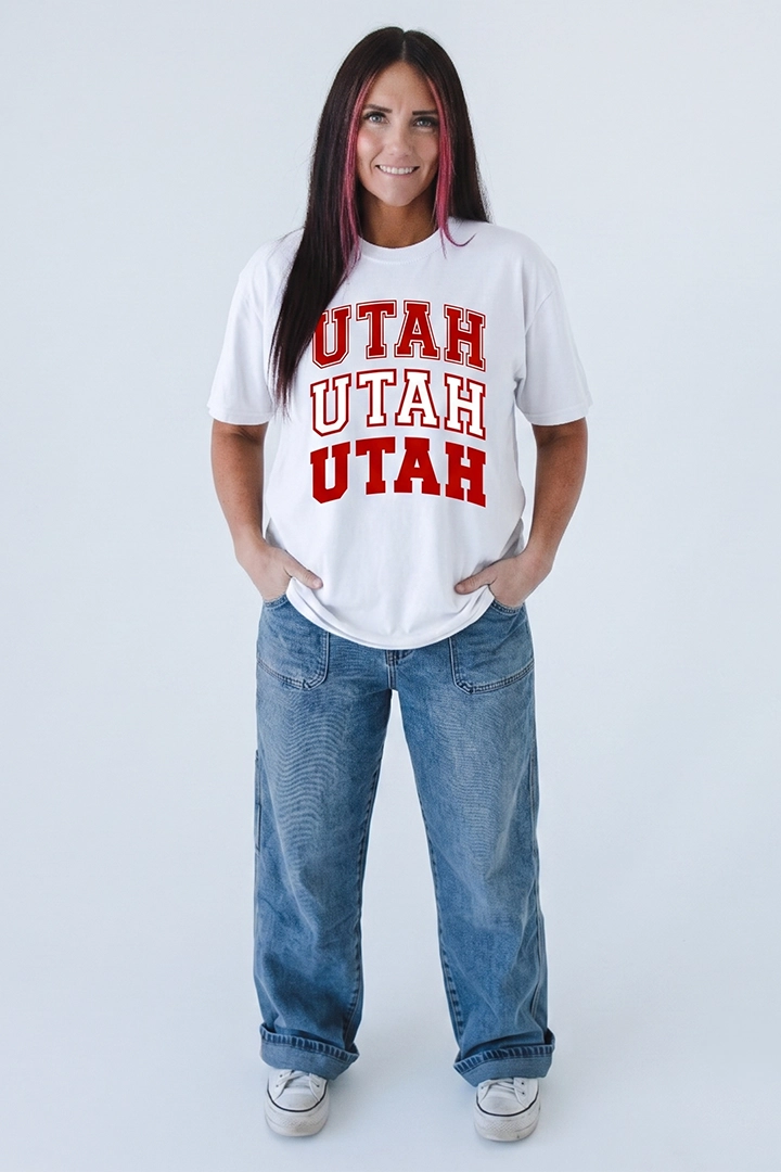 A white short-sleeved t-shirt. The front of the shirt features the word 'UTAH' stacked vertically three times in a bold, arched collegiate font. The top word is outlined in red, the middle word is white with a red outline, and the bottom word is solid red.