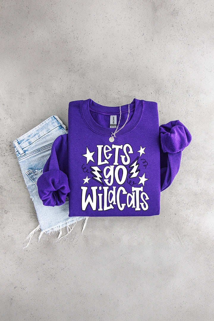 Flat lay of a bright purple crewneck sweatshirt printed with white text reading "Lets Go Wildcats" surrounded by stars and lightning bolts, styled with distressed light-wash denim shorts and a silver pendant necklace on a grey concrete background.