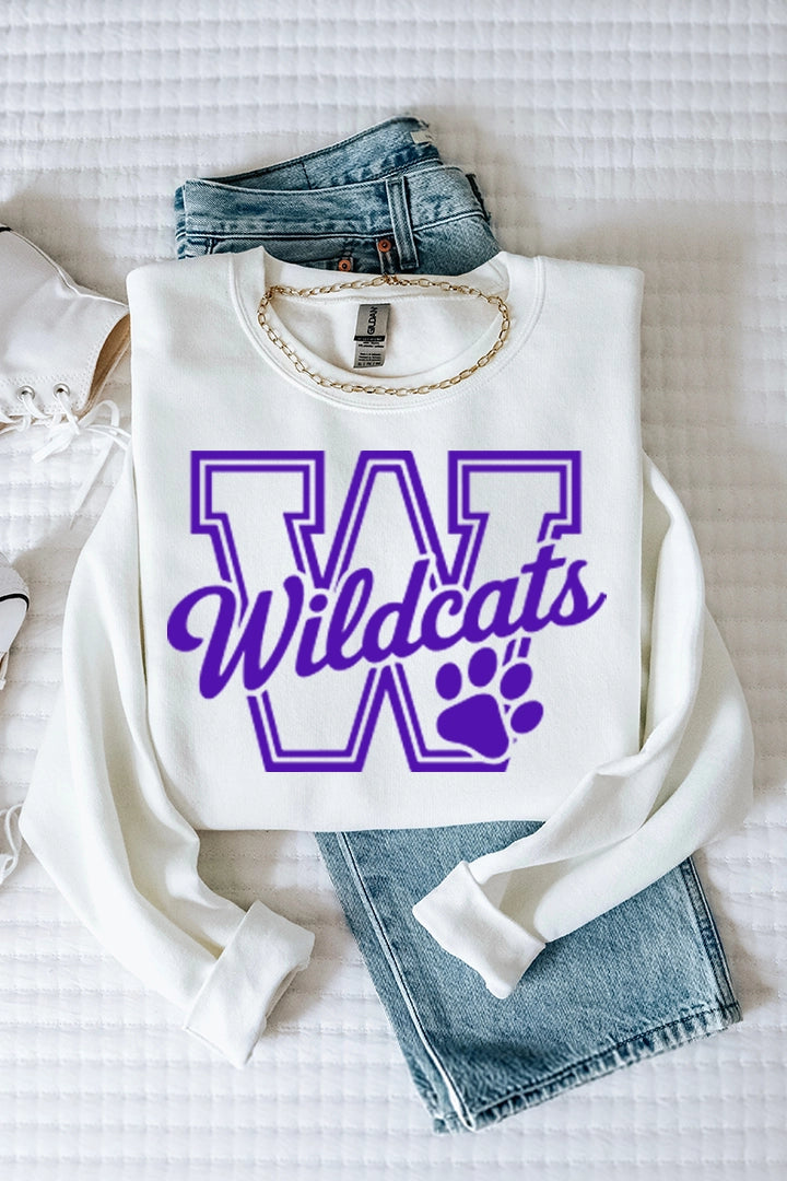 Flat lay view of a white crewneck sweatshirt styled on a white textured blanket with medium-wash blue jeans, a gold chain necklace, and white high-top sneakers. The sweatshirt features a large purple outlined block letter "W", script text reading "Wildcats", and a purple paw print graphic.