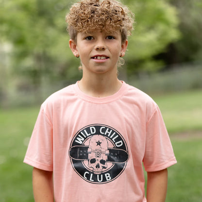 A young boy with blonde curly hair and smile stands with his hands in his pockets, wearing a light peach short-sleeved graphic apparel t-shirt with a circular black and white 'Wild Child Club' graphic. The graphic includes a skull with a star, wings, and the text 'WILD CHILD CLUB'. He is also wearing blue denim jeans with distressed details and is standing on a green lawn with blurred trees in the background.