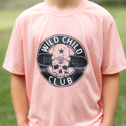 A young boy with blonde curly hair and smile stands with his hands in his pockets, wearing a light peach short-sleeved graphic apparel t-shirt with a circular black and white 'Wild Child Club' graphic. The graphic includes a skull with a star, wings, and the text 'WILD CHILD CLUB'. He is also wearing blue denim jeans with distressed details and is standing on a green lawn with blurred trees in the background.