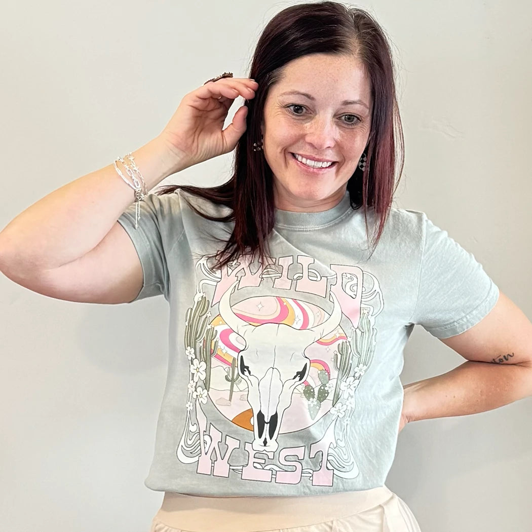 A fair-skinned person with shoulder-length dark hair smiles, wearing a womens graphic tshirts, light green graphic apparel t-shirt with a stylized graphic in muted pinks, oranges, and greens. The graphic includes a cow skull with desert elements like cacti and a wavy landscape behind it, and the text "WILD" above and "WEST" below the central image. The design is western style.