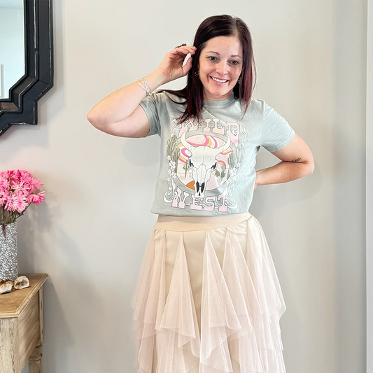 A fair-skinned person with shoulder-length dark hair smiles, wearing a womens graphic tshirts, light green graphic apparel t-shirt with a stylized graphic in muted pinks, oranges, and greens. The graphic includes a cow skull with desert elements like cacti and a wavy landscape behind it, and the text "WILD" above and "WEST" below the central image. The design is western style.