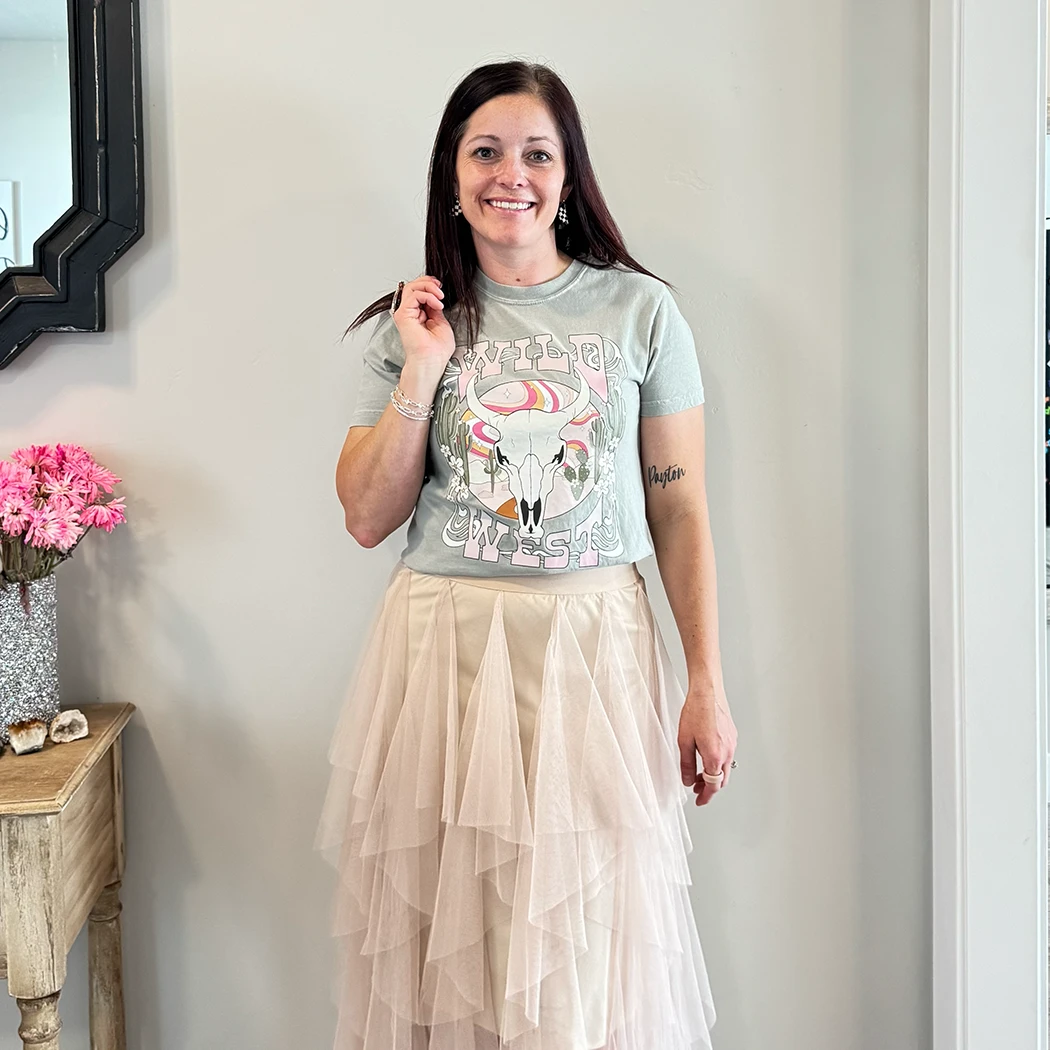 A fair-skinned person with shoulder-length dark hair smiles, wearing a womens graphic tshirts, light green graphic apparel t-shirt with a stylized graphic in muted pinks, oranges, and greens. The graphic includes a cow skull with desert elements like cacti and a wavy landscape behind it, and the text "WILD" above and "WEST" below the central image. The design is western style.