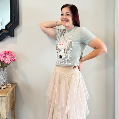 A fair-skinned person with shoulder-length dark hair smiles, wearing a womens graphic tshirts, light green graphic apparel t-shirt with a stylized graphic in muted pinks, oranges, and greens. The graphic includes a cow skull with desert elements like cacti and a wavy landscape behind it, and the text "WILD" above and "WEST" below the central image. The design is western style.