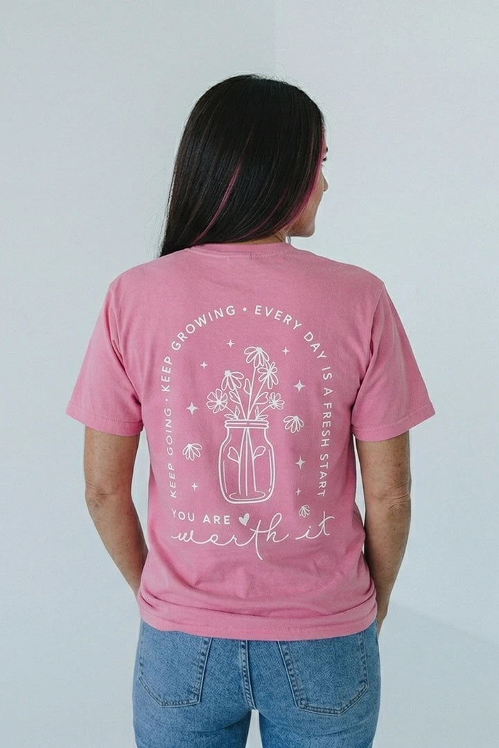 rose-pink short-sleeved t-shirt. The back of the shirt features a white line-art graphic of flowers in a jar, arched text that reads 'KEEP GOING • KEEP GROWING • EVERY DAY IS A FRESH START,' and 'YOU ARE worth it' in a mix of block and script fonts at the bottom."