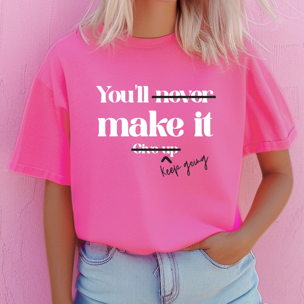 Neon pink graphic shirt with white text that says You'll never make it. Give up. The never is crossed out and the give up is crossed out with keep going written next to it. 