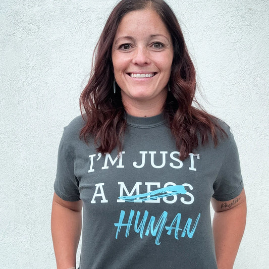 A person stands in front of a white wall, wearing a dark gray t-shirt with a white and blue graphic. The text reads "I'M JUST A MESS HUMAN," with the word "mess" having a blue line drawn through it and "human" written in blue cursive underneath.