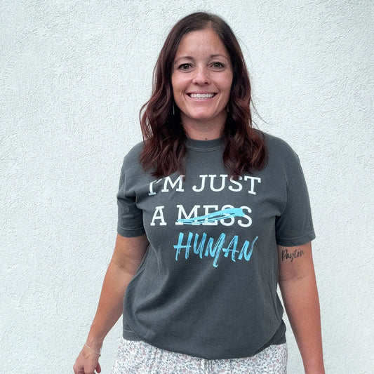 A person stands in front of a white wall, wearing a dark gray t-shirt with a white and blue graphic. The text reads "I'M JUST A MESS HUMAN," with the word "mess" having a blue line drawn through it and "human" written in blue cursive underneath.
