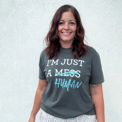 A person stands in front of a white wall, wearing a dark gray t-shirt with a white and blue graphic. The text reads "I'M JUST A MESS HUMAN," with the word "mess" having a blue line drawn through it and "human" written in blue cursive underneath.