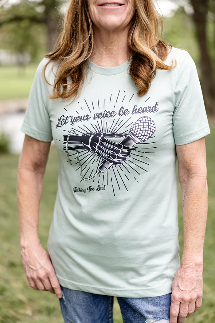 Heather dust shirt with positive message that says Let Your Voice Be Heard. The design has a Skelton hand with a microphone.  