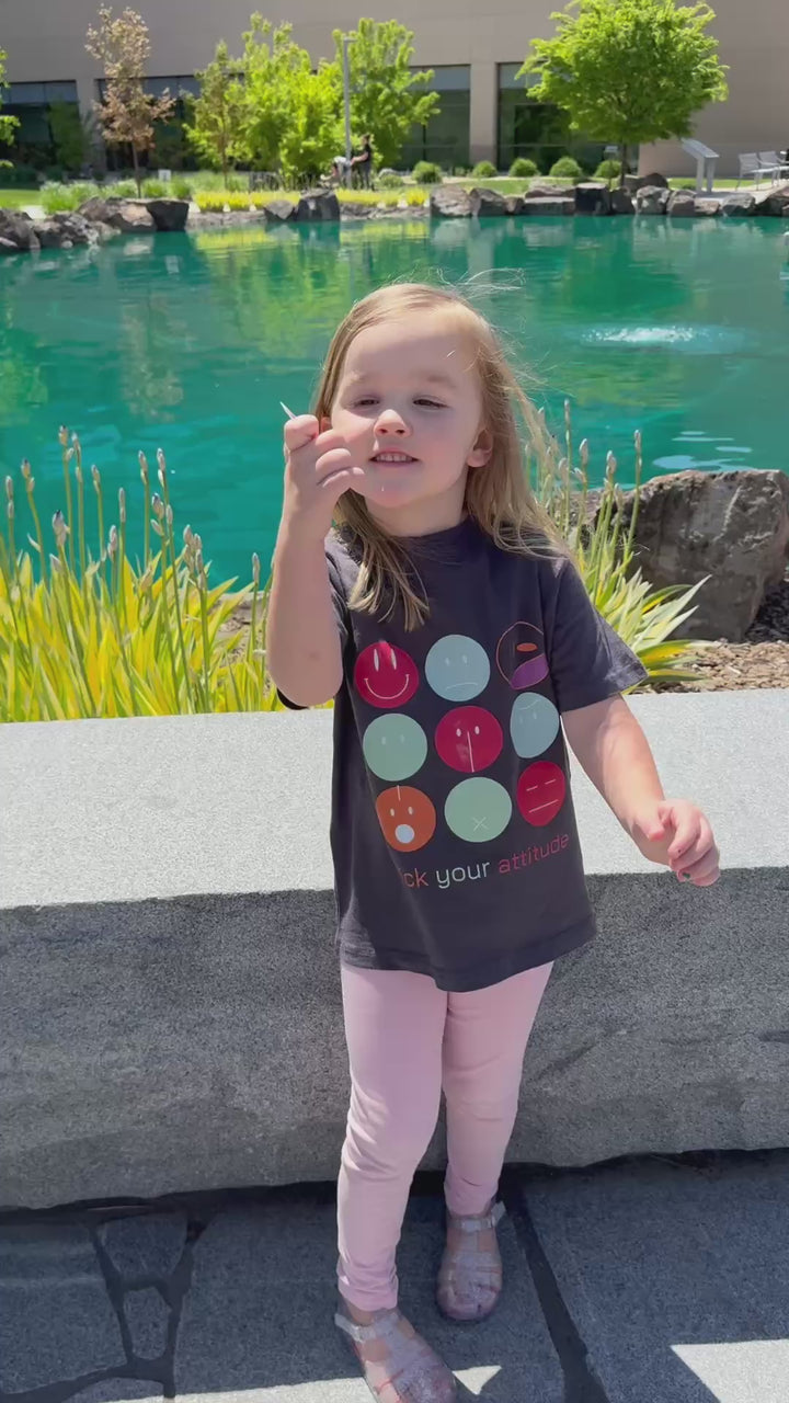 A young child with blonde hair smiles while standing outdoors in front of a pond and modern buildings. The child is wearing a dark gray graphic apparel t-shirt with a grid of colorful smiley faces and the text "Pick your attitude" printed on it, along with light pink leggings and gray shoes.