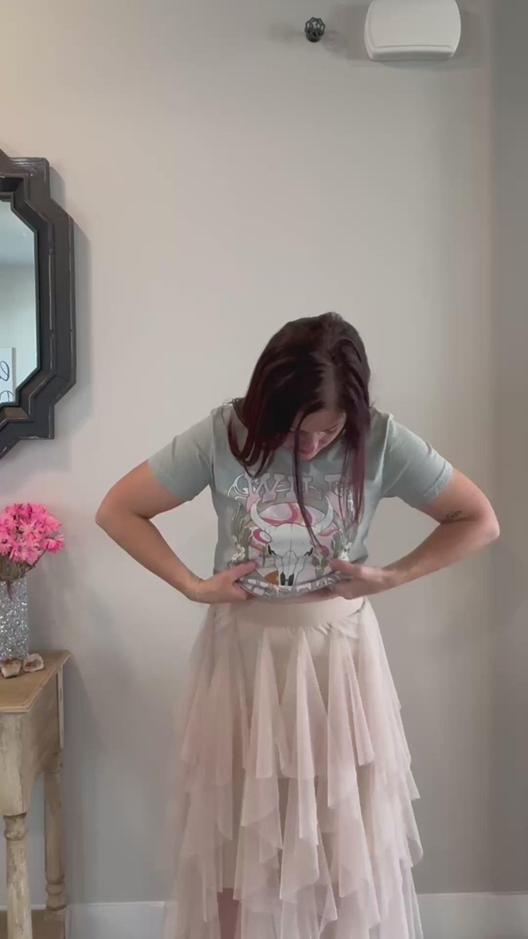 A fair-skinned person with shoulder-length dark hair smiles, wearing a womens graphic tshirts, light green graphic apparel t-shirt with a stylized graphic in muted pinks, oranges, and greens. The graphic includes a cow skull with desert elements like cacti and a wavy landscape behind it, and the text "WILD" above and "WEST" below the central image. The design is western style.